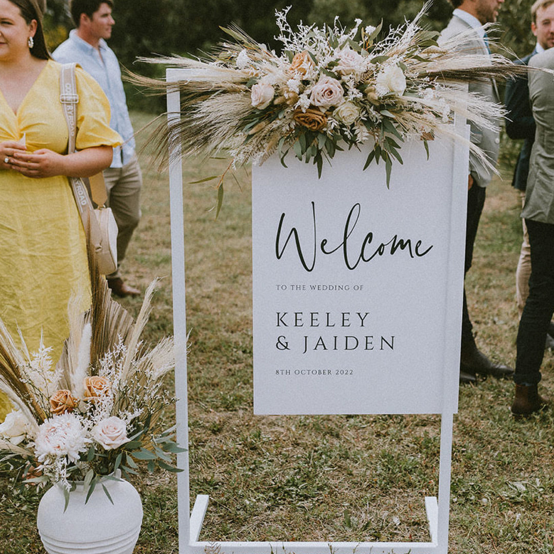 Modern wedding welcome sign with floral arrangement on top.