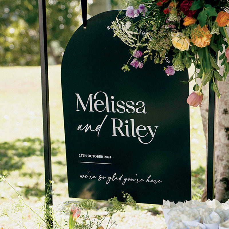 Modern wedding welcome sign with floral arrangement on top.