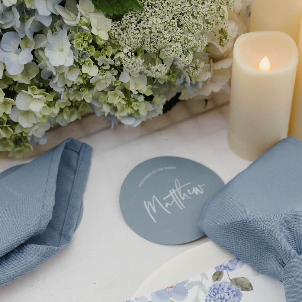 Billie round wedding place card in dusty blue with modern script styling, displayed with blue napkins and floral table décor.