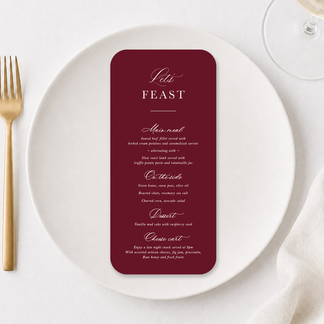 Burgundy wedding menu with elegant calligraphy on a white dinner plate with gold cutlery